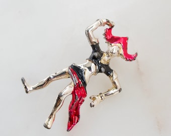 Puede incluir: Un broche plateado que representa a una bailarina estilizada con un traje rojo y negro. La bailarina lleva un sombrero rojo y sostiene una bufanda roja.