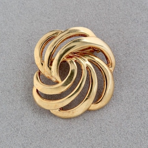 May include: A gold-colored brooch with a swirling, abstract design. The brooch features multiple curved, overlapping elements, creating a three-dimensional effect. The jewelry piece is set against a gray background.
