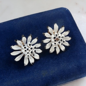 Puede incluir: Un par de pendientes blancos en forma de flor con centros negros. Los pendientes están sobre un fondo de terciopelo azul.