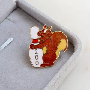 Op de afbeelding: Een gouden speld met een cartoon-eekhoorn die een witte bowlingpin omarmt met een rode streep en het nummer "200" erop gedrukt.