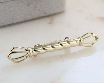 May include: A gold-toned twisted metal brooch with two decorative ends. The brooch is on a white surface.