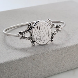 May include: A silver-toned bangle bracelet featuring a central oval locket with an intricate, engraved script monogram. The locket is flanked by ornate, scroll-like metalwork detailing that connects to the thin, polished circular band.