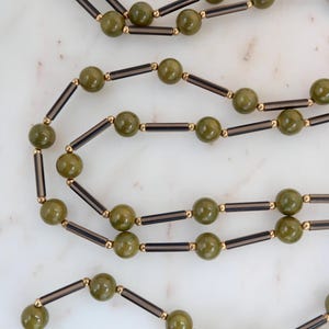 Puede incluir: Collares de cuentas verde oliva con detalles en negro y dorado. Los collares presentan cuentas redondas intercaladas con cuentas cilíndricas negras y conectores dorados. Las joyas se exhiben sobre una superficie de mármol blanco.