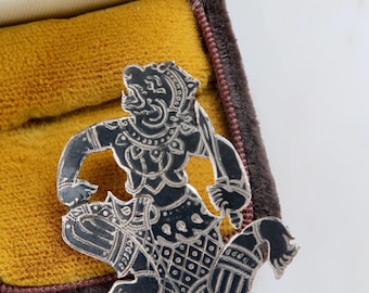 May include: A silver brooch in the shape of a mythical creature with intricate details. The brooch is sitting in a velvet box.