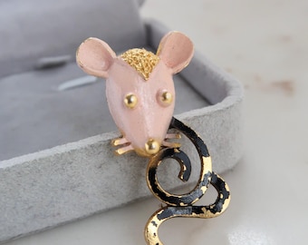 May include: A pink and gold mouse brooch with a black and gold swirled tail. The mouse has gold accents on its ears, eyes, and whiskers. The brooch is set against a gray background.