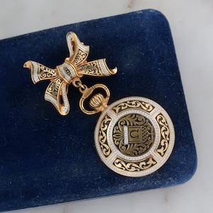 May include: A gold-tone brooch with a black and white enamel design. The brooch features a bow with a pendant in the shape of a circle with a monogram.