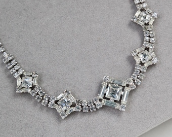 May include: A silver-toned necklace with clear, square-cut and round-cut rhinestones. The necklace features a series of square-cut stones set in a geometric pattern, connected by smaller, round rhinestones. The jewelry is displayed on a gray surface.