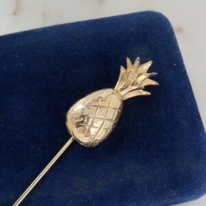 Op de afbeelding: Een goudkleurige ananasvormige speld met een lange, slanke pin. De ananas heeft een getextureerd oppervlak en is geplaatst tegen een donkerblauwe achtergrond. De speld is waarschijnlijk een decoratief accessoire.