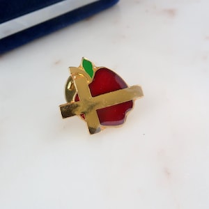 May include: A gold-toned pin with a red apple design. The apple has a green leaf and is crossed by a gold band.