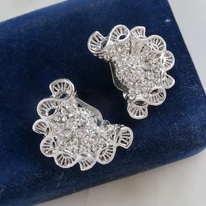 May include: A pair of silver clip-on earrings with a delicate, ornate design. The earrings are covered in sparkling clear rhinestones.