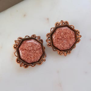 May include: A pair of copper-toned earrings with a glittery, reddish-brown stone set in a decorative, octagonal frame.