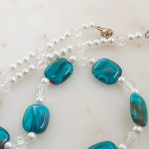 May include: A necklace featuring a combination of white faux pearls, clear crystal beads, and vibrant teal and blue glass beads. The necklace has a silver clasp and is displayed on a white marble surface.