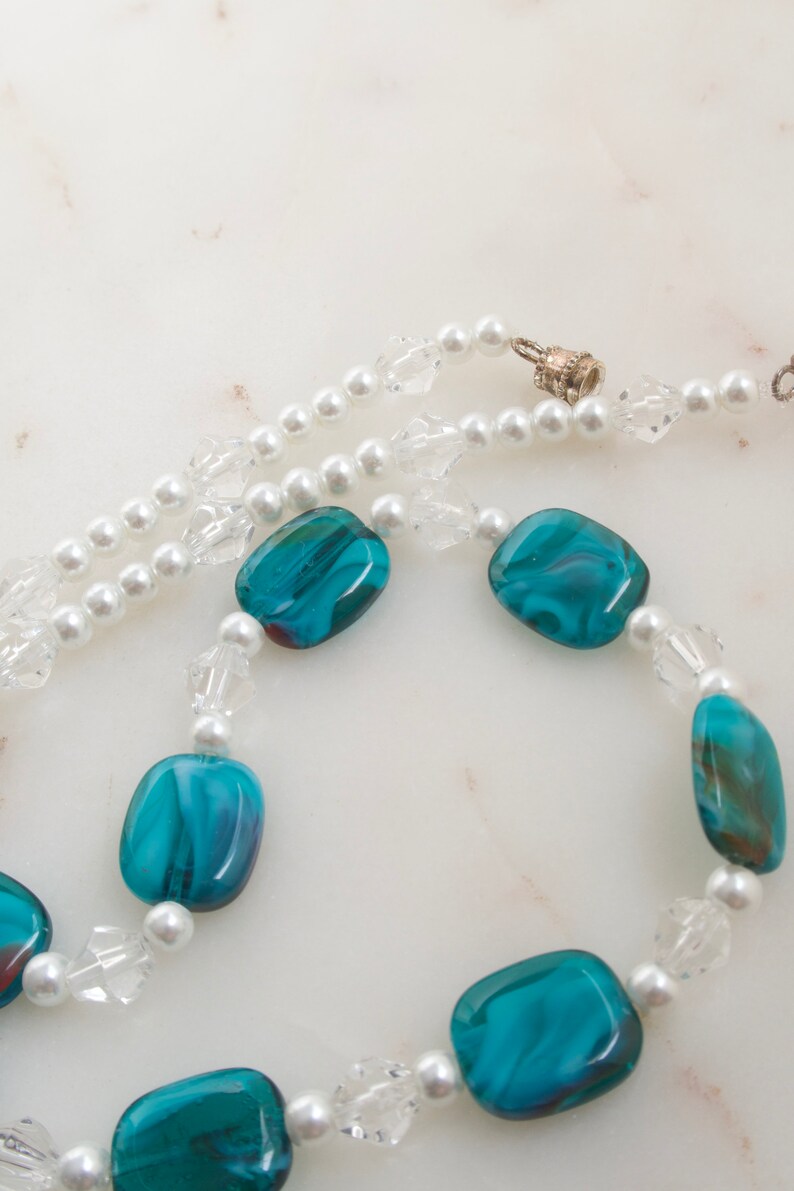 May include: A necklace featuring a combination of white faux pearls, clear crystal beads, and vibrant teal and blue glass beads. The necklace has a silver clasp and is displayed on a white marble surface.