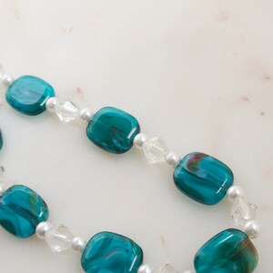May include: A necklace featuring teal and clear beads. The teal beads are rectangular and have a marbled appearance. The clear beads are faceted and interspersed with small, round white beads. The necklace is displayed on a white surface.