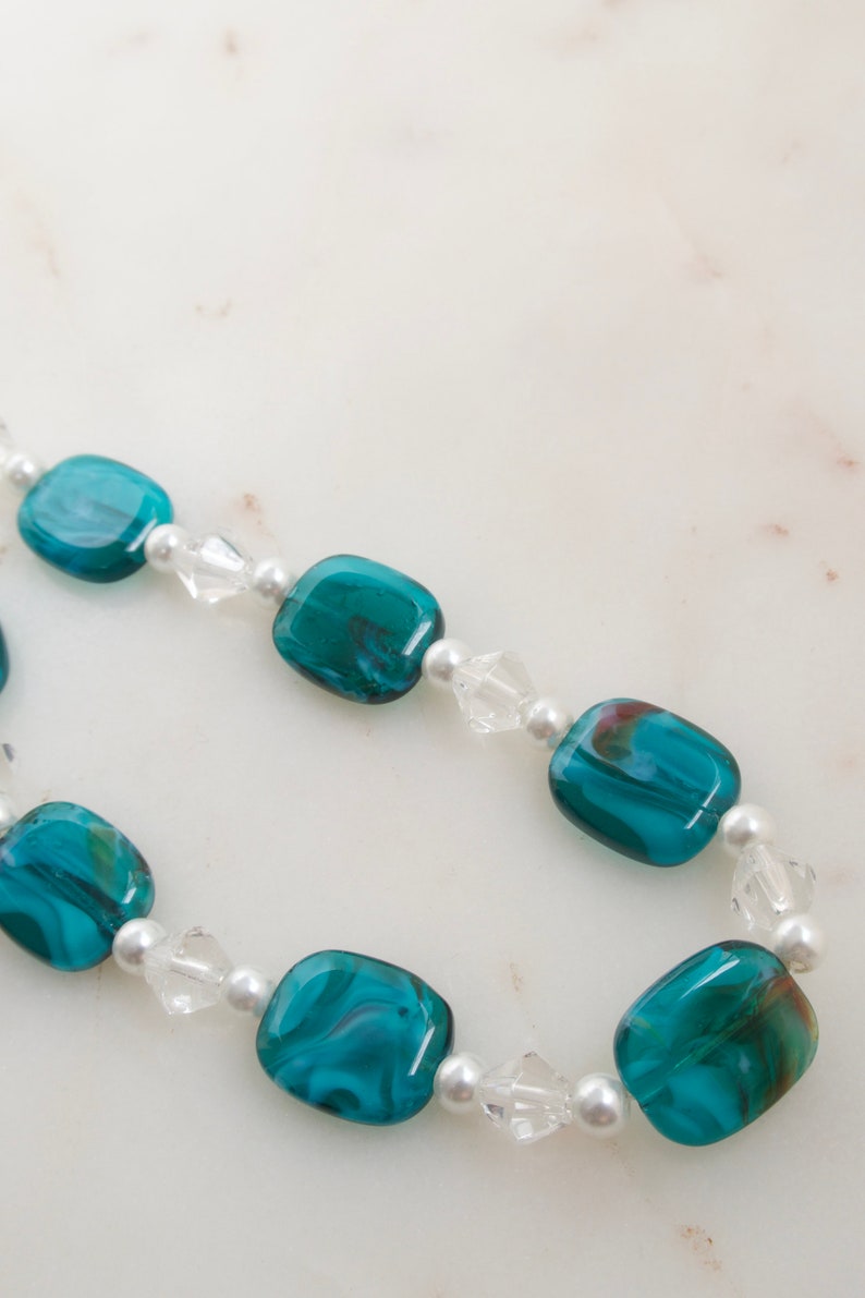 May include: A necklace featuring teal and clear beads. The teal beads are rectangular and have a marbled appearance. The clear beads are faceted and interspersed with small, round white beads. The necklace is displayed on a white surface.