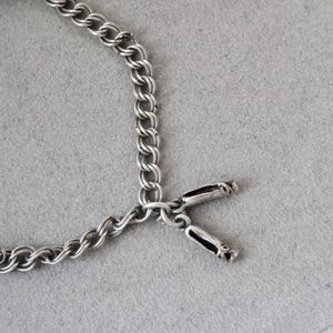 May include: Close-up of a silver chain bracelet with a toggle clasp. The bracelet features a double-link chain design. The toggle clasp has a rectangular shape with black accents. The bracelet is displayed on a grey surface.