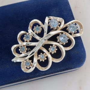Puede incluir: Broche vintage plateado con diseño floral. El broche presenta formas arremolinadas en forma de cinta, adornadas con pedrería azul claro. El broche está sobre una superficie de terciopelo azul oscuro.