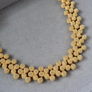 Könnte beinhalten: Eine goldene Halskette mit einer Kette aus kleinen, strukturierten Kugeln. Die Halskette wird auf einem grauen Samtstoff präsentiert.