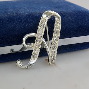 May include: Silver-toned letter "A" brooch, encrusted with clear rhinestones. The brooch is set against a blue velvet surface, with a silver bar pin on the back. The brooch is a decorative accessory.