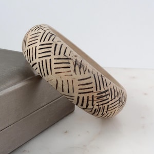 May include: A wooden bangle with a black and white geometric pattern. The bangle is light brown and has a smooth surface.