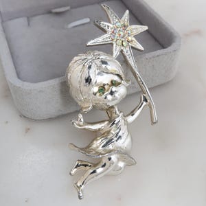 May include: Silver-toned brooch featuring a whimsical fairy design. The fairy holds a star-shaped wand with iridescent rhinestones. The brooch has green gem eyes and is set against a gray velvet jewelry box.