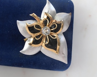 May include: A silver and gold tone brooch with a black enamel flower and a clear rhinestone in the center.