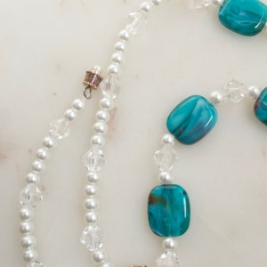 May include: A necklace featuring white faux pearls, clear crystal beads, and rectangular turquoise-colored glass beads. The necklace has a magnetic clasp and is displayed on a white marble surface.