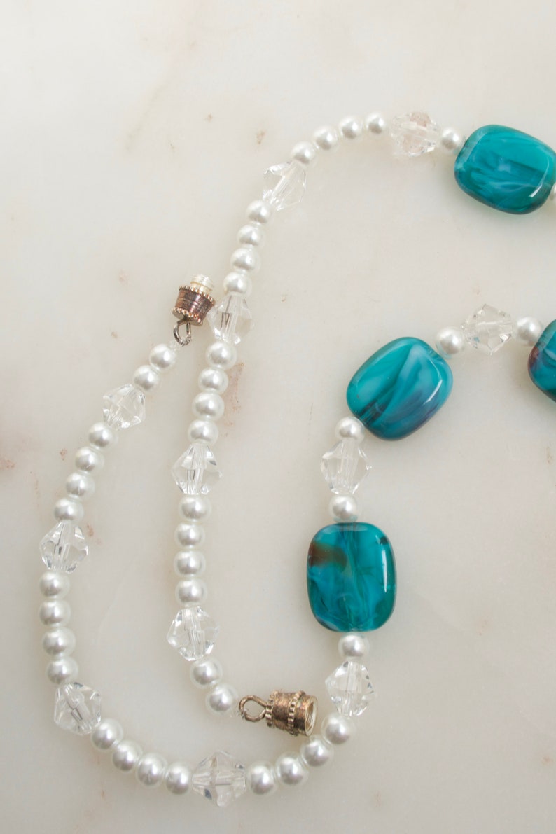 May include: A necklace featuring white faux pearls, clear crystal beads, and rectangular turquoise-colored glass beads. The necklace has a magnetic clasp and is displayed on a white marble surface.