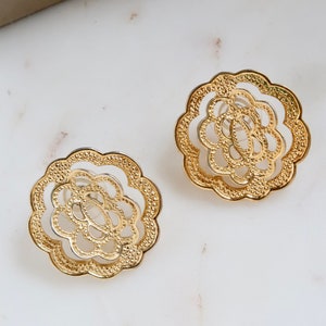 May include: A pair of gold-tone earrings with a delicate floral design. The earrings are made of metal and have a circular shape.