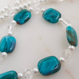 May include: A necklace featuring large, teal-colored, rectangular beads with a marbled effect, interspersed with small, white faux pearls and clear crystal beads. The necklace has a silver clasp and a strand of white faux pearls.