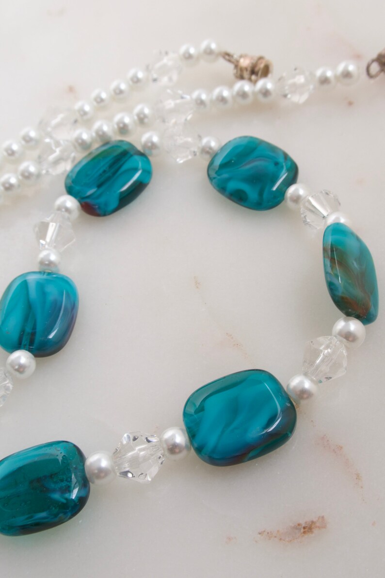 May include: A necklace featuring large, teal-colored, rectangular beads with a marbled effect, interspersed with small, white faux pearls and clear crystal beads. The necklace has a silver clasp and a strand of white faux pearls.