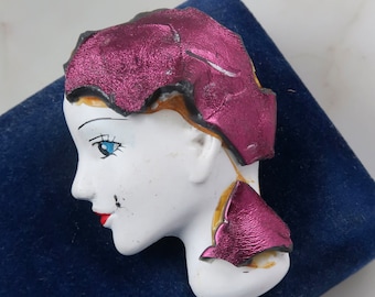Puede incluir: Un broche de cerámica blanca que representa la cara de una mujer con un cubrecabezas rosa y dorado. La mujer tiene ojos azules y labios rojos.