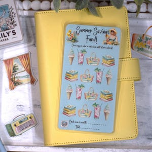 May include: A light blue rectangular savings tracker sticker sheet featuring the text Summer Savings Fund! and illustrations of ice cream cones, cocktails, beach bags, and books, placed on a yellow notebook with additional stickers nearby.