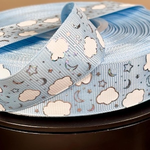 May include: A spool of light blue ribbon with a repeating pattern of white clouds, stars, and crescent moons. The ribbon is rolled up and displayed on a black surface. The ribbon is approximately 1 inch wide.