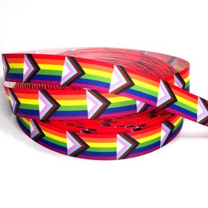 Op de afbeelding: Een rol grosgrain lint met een herhalend patroon van de Progress Pride vlag. Het ontwerp bestaat uit horizontale strepen in rood, oranje, geel, groen, blauw en violet, geaccentueerd door een chevronvorm in wit, roze, lichtblauw, bruin en zwart.