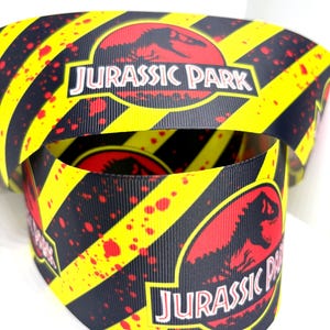 Peut inclure: Gros plan sur un ruban avec le logo "Jurassic Park". Le ruban présente un motif de rayures noires et jaunes, avec des éclaboussures rouges. Le logo est un ovale rouge avec une silhouette de dinosaure et le texte "JURASSIC PARK".