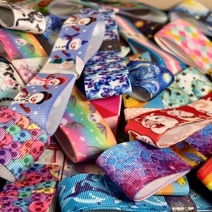 Op de afbeelding: Een verzameling kleurrijke linten met diverse patronen. Ontwerpen omvatten sneeuwmannen, schedels, vleermuizen en bloemmotieven. De linten zijn in tinten blauw, roze, paars en rood, met witte en bedrukte randen. Ideaal voor knutselen en cadeauverpakking.