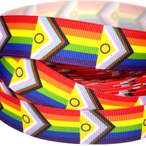 Könnte beinhalten: Eine Rolle Ripsband mit einem sich wiederholenden Muster der Progress Pride Flagge. Das Design zeigt horizontale Regenbogenstreifen, die von einer Chevron-Form in Braun, Schwarz, Hellblau, Pink und Weiß mit einem violetten Kreis durchbrochen werden.