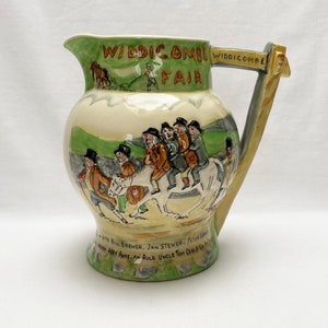 May include: A ceramic jug with a green and white design depicting a group of people riding horses. The jug has a handle and the text "WIDDICOMBE FAIR" is printed on the front.