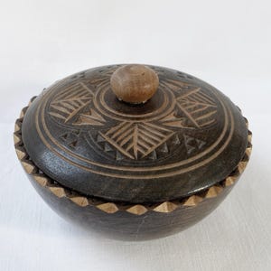 May include: A dark brown wooden bowl with a lid. The lid has carved geometric patterns and a small wooden knob. The bowl's rim has a decorative, notched design. The bowl is likely used for storage or display.