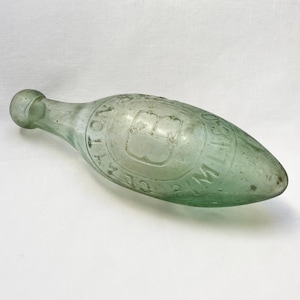 Puede incluir: Una botella de vidrio verde con fondo redondeado y cuello largo y estrecho. La botella tiene un diseño circular en relieve con el texto "WILSON'S" y un logotipo de corona.