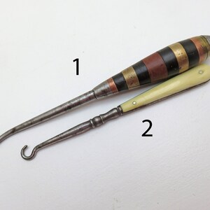May include: Two antique shoe horns with different handles. The first shoe horn has a black, copper and gold striped handle. The second shoe horn has a white handle.