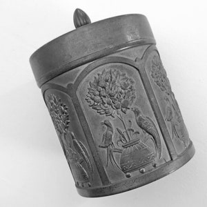 May include: A silver metal container with a lid, featuring an embossed design of birds and flowers. The container has a rounded shape and a smooth surface.