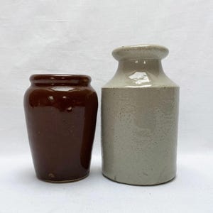 May include: Two antique stoneware jars. One is a dark brown, glazed jar. The other is a light grey jar with a rounded top. Both jars are on a white background.