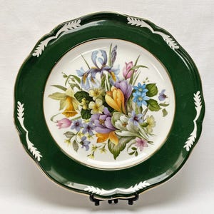 May include: A white ceramic dinner plate with a green rim and a floral design featuring a bouquet of irises, crocuses, and other flowers. The plate has a gold trim.