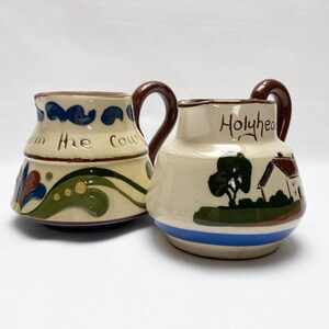 May include: Two ceramic jugs with brown handles. One is cream-coloured with blue and green floral designs and the text "The Cow". The other is cream-coloured with a blue stripe, a house and tree design, and the text "Holyhead".