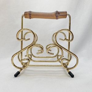 May include: Gold metal napkin holder with a bamboo handle. The holder has a decorative, ornate design.