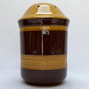 May include: A ceramic salt shaker with a brown body and a tan top. The shaker has a decorative band of small circles around the middle. The top has three holes for dispensing salt. A vintage kitchenware item.