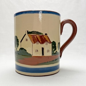 May include: A ceramic mug with a hand-painted cottage scene. The mug has a cream base, a brown handle, and a blue rim. The cottage has a red roof and is surrounded by green trees. The mug is approximately 10 cm tall.