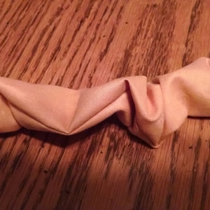 May include: A close-up of a light peach-colored fabric headband, folded and twisted into a decorative shape.